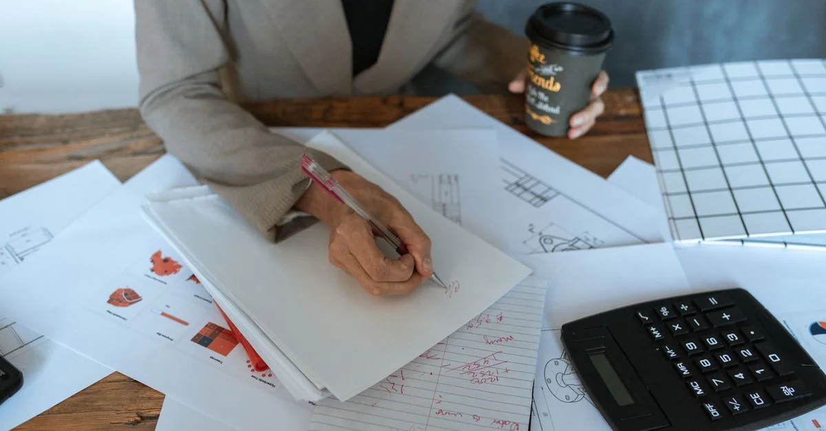 Gros plan sur une personne écrivant à un bureau avec des documents et un café, symbolisant la productivité.